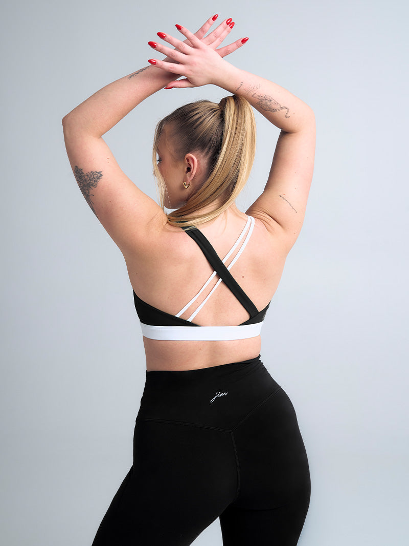 Tył stanika JOYINME Empower w czerni z unikalnym systemem ramiączek. Szeroki czarny pas krzyżuje się z delikatnymi białymi ramiączkami, tworząc geometryczny wzór na plecach. Stanik sfotografowany podczas dynamicznego ruchu, ukazujący funkcjonalność i elastyczność materiału.